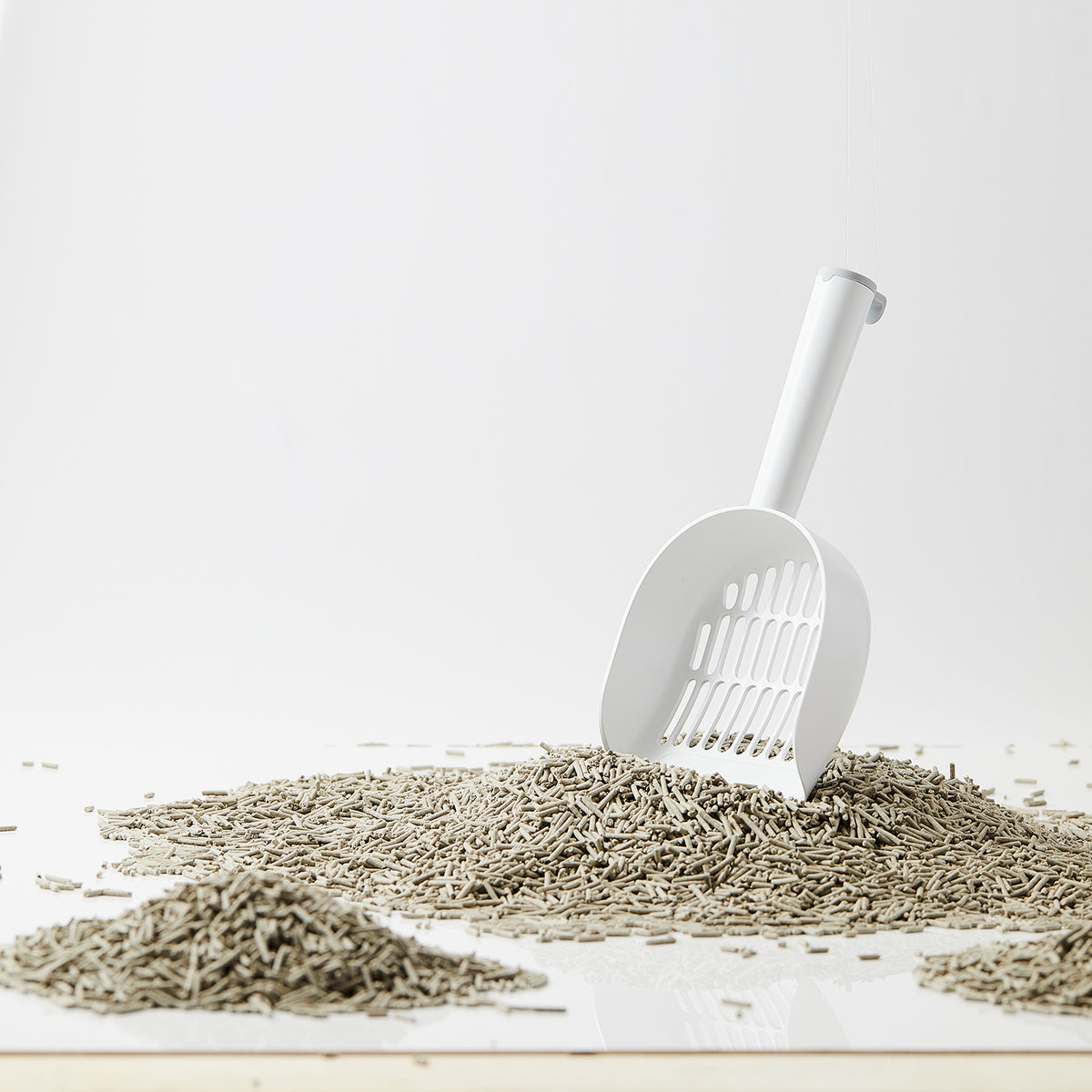 A white litter scoop resting on a pile of gray cat litter, with additional scattered litter on a clean white surface against a light gray background.