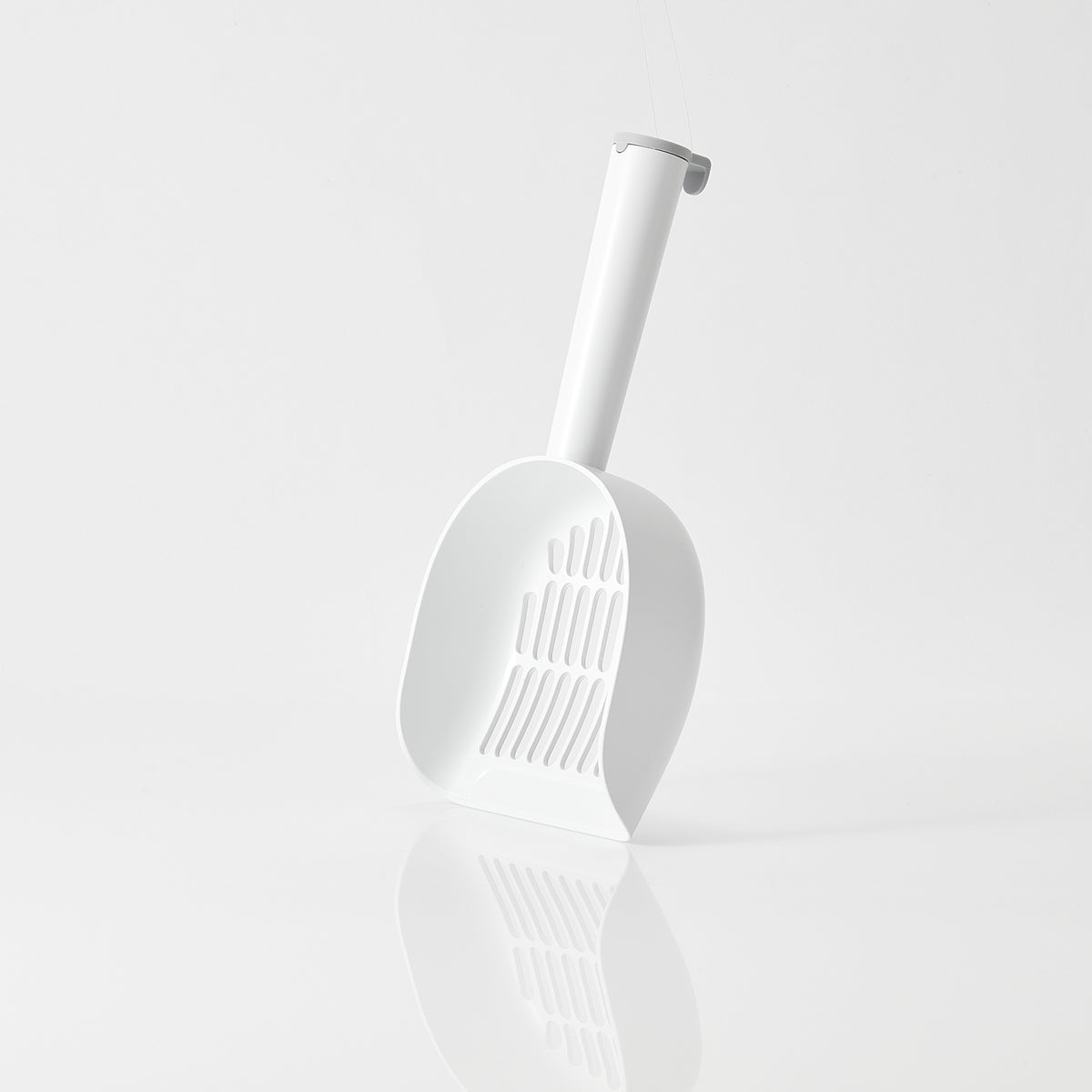 A white plastic cat litter scoop with a slotted front and a curved handle, shown standing upright on a clean white reflective surface against a light gray background.